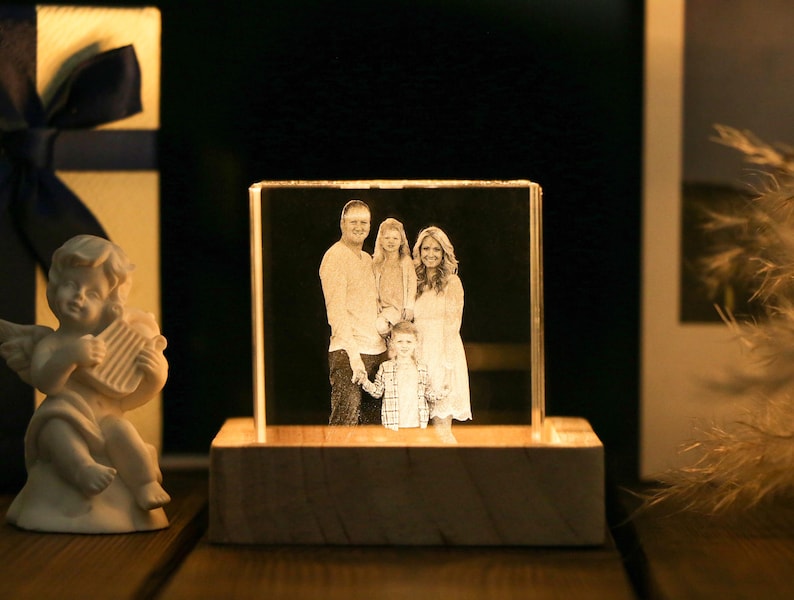 May include: A family portrait is displayed within a clear, rectangular crystal block, illuminated from below by a wooden base. The image shows a family of four. A small, white cherub statue with a harp is on the left, and a gift box is in the background.