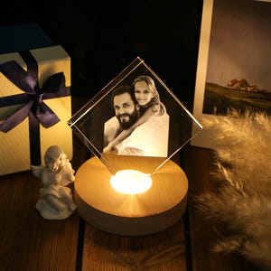 May include: A lit, diamond-shaped crystal photo frame displaying a black and white image of a man and a girl. The frame sits on a wooden base with a warm light. A gift box with a navy blue ribbon and a small angel figurine are also present.