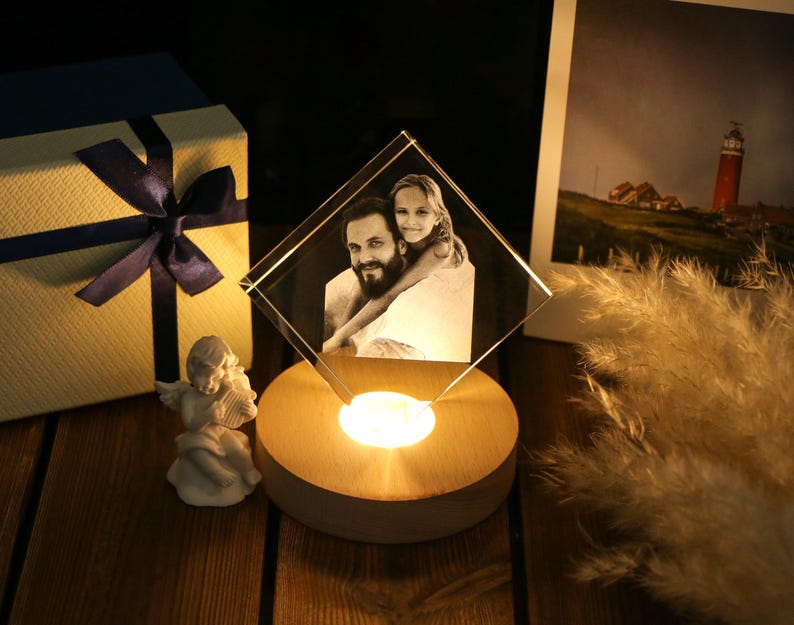 May include: A lit, diamond-shaped crystal photo frame displaying a black and white image of a man and a girl. The frame sits on a wooden base with a warm light. A gift box with a navy blue ribbon and a small angel figurine are also present.