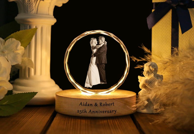 May include: A lighted, crystal-like sphere with a couple embracing inside, set on a wooden base. The base is engraved with "Aidan & Robert 25th Anniversary." A decorative angel statue and a gift box are in the background.