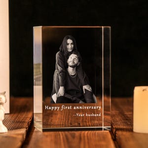 Może przedstawiać: Przezroczysty, prostokątny blok kryształowy ze zdjęciem pary w czerni i bieli. Na przodzie nadrukowano tekst "Happy first anniversary --Your husband". Świeca i oprawione zdjęcie znajdują się w tle.
