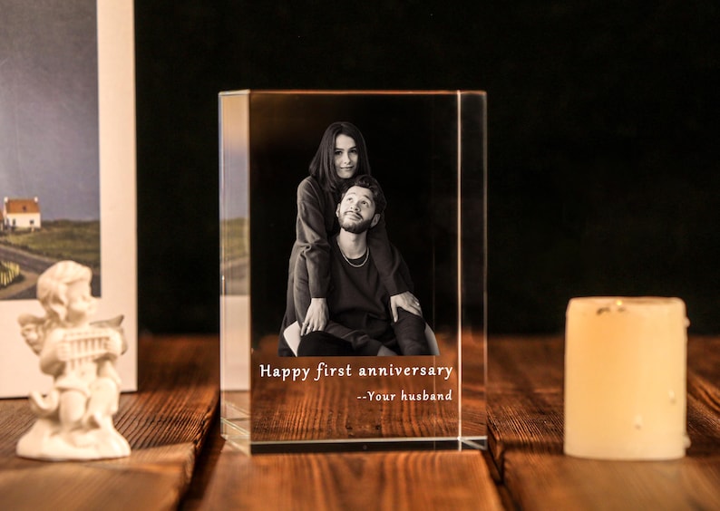Może przedstawiać: Przezroczysty, prostokątny blok kryształowy ze zdjęciem pary w czerni i bieli. Na przodzie nadrukowano tekst "Happy first anniversary --Your husband". Świeca i oprawione zdjęcie znajdują się w tle.