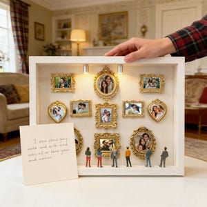 Op de afbeelding: Een witte schaduwdoos met meerdere kleine, sierlijke gouden fotolijsten, elk met een foto. Miniatuurfiguren staan onder de lijsten. Een handgeschreven notitie staat ervoor.