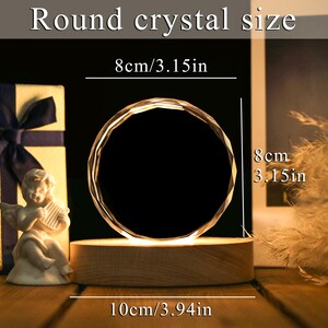May include: A round crystal with a faceted design, illuminated from below, sits on a wooden base. The crystal is 3.15 inches in diameter and 3.94 inches wide. The text "Round crystal size" is at the top. A small angel statue and a gift box are in the background.