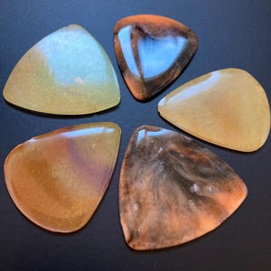 Può includere: Cinque plettri per chitarra in vari colori, tra cui oro, marrone e arancione, disposti su una superficie scura. I plettri hanno un aspetto lucido e traslucido, con motivi e vortici unici.