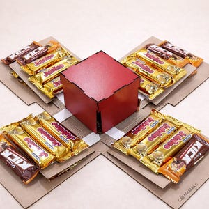 Peut inclure: Une boîte cadeau carrée rouge avec un design découpé au laser, entourée de quatre plateformes en carton. Chaque plateforme contient plusieurs barres chocolatées dans des emballages dorés et bruns.