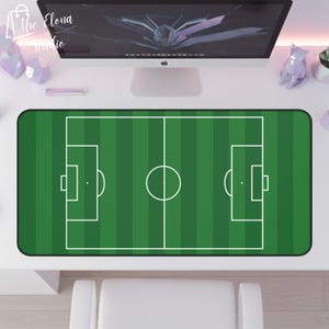 May include: A rectangular green desk mat featuring a soccer field design with white lines. The mat has rounded corners and is placed on a white desk. The Elona Studio logo is visible in the top left corner.