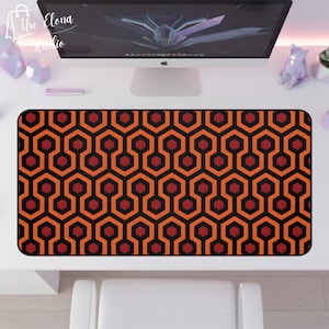 May include: A rectangular desk mat featuring a repeating geometric pattern in black, orange, and red. The design consists of interconnected hexagons and elongated shapes. The mat is on a white desk, with a computer monitor and other desk accessories visible in the background.