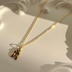 Op de afbeelding: Een goudkleurige ketting met een kleine hanger in de vorm van een paar balletschoenen. De hanger hangt aan een fijne, dunne goudkleurige ketting en reflecteert het licht met een gepolijste metalen afwerking en gladde, gebogen vormen.