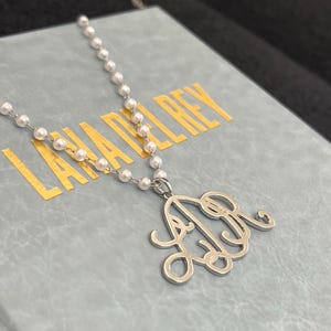 May include: A silver monogram necklace with a pearl chain. The pendant features an ornate script design of the letters L and R. The necklace rests on a light blue surface with the gold text "LANA DEL REY".