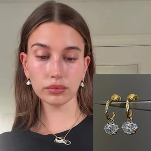 Peut inclure: Une paire de boucles d'oreilles créoles en or avec une pierre précieuse transparente de taille ovale suspendue. Les boucles sont présentées portées aux lobes et tenues par une pince, révélant la finition en or poli et les pierres blanches facettées.