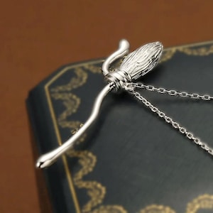 Puede incluir: Un collar con colgante de escoba plateada. El colgante presenta un diseño detallado de escoba con un mango curvado y una cabeza texturizada. El collar tiene una cadena delicada.