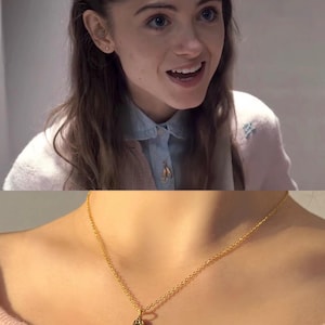 Peut inclure: Un collier en métal doré composé d'une chaîne fine et d'un pendentif en forme de paire de chaussons de danse. Le pendentif est suspendu au centre de la chaîne, qui repose contre la zone de la clavicule, mettant en valeur une finition métallique.