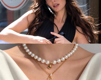 May include: A layered necklace set featuring a strand of white pearls with a small gold teardrop pendant and a gold chain necklace with a toggle clasp and a small clear stone accent, worn against a white fabric garment.