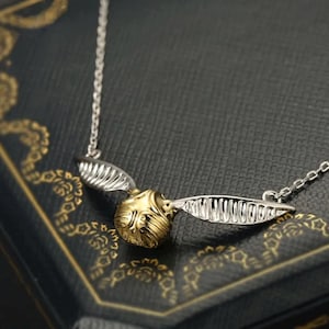 Puede incluir: Un collar plateado con un colgante de Snitch dorado y alas plateadas. El colgante presenta detalles texturizados y cuelga de una fina cadena. El collar se presenta sobre una superficie oscura estampada.