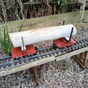 May include: A model railway scene featuring a light-coloured log on two red flatbed wagons. The wagons are on a model railway track, supported by a wooden trestle. The background includes grass and foliage.