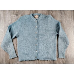 Könnte beinhalten: Hellblauer Cardigan mit Rundhalsausschnitt und langen Ärmeln. Der flauschig strukturierte Strickpullover ist mit silberfarbenen Knöpfen versehen. Ein kleines Etikett ist am Ausschnitt sichtbar.