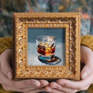May include: A small, square, gold-framed painting of a whiskey glass filled with ice and amber liquid. The painting is held in two hands, and the frame has ornate detailing.