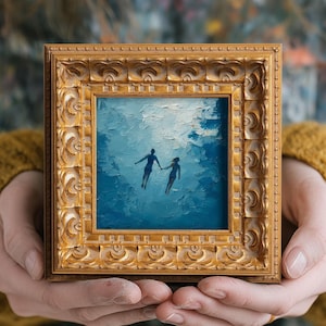 Puede incluir: Una pequeña pintura cuadrada en un marco dorado ornamentado representa dos figuras silueteadas tomadas de la mano en una escena submarina azul texturizada. El marco lo sostiene una persona que lleva un jersey amarillo.