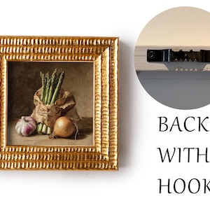 May include: A framed still life painting featuring asparagus, garlic, and an onion. The artwork is in a gold ornate frame. The back of the frame has a hook for hanging. The painting is a still life of fresh vegetables.