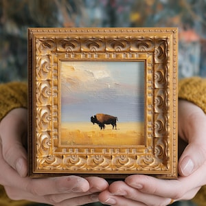 May include: A small, square painting of a bison on a prairie under a cloudy sky. The artwork is in a gold, ornate frame. The painting is held in a person's hands, with a yellow sweater in the background.