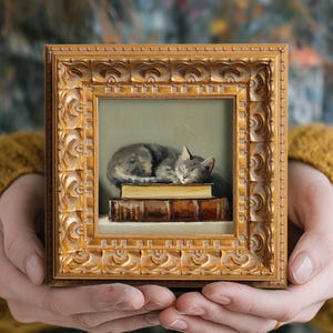 May include: A framed painting of a sleeping gray cat resting on a stack of books. The artwork is in a gold ornate frame. The cat is curled up, with its eyes closed. The books are brown with gold lettering.