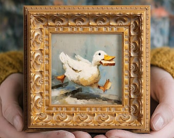 May include: A small, square, gold-framed painting depicts a white duck with a piece of bread in its beak. The duck is running on a light blue background. The frame has an ornate, carved design.