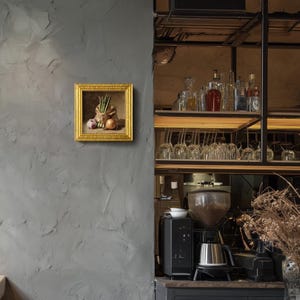 May include: A framed still life painting with a gold frame. The painting depicts a burlap sack filled with asparagus, an onion, and a garlic bulb. The painting is mounted on a textured gray wall, with a bar and coffee station in the background.