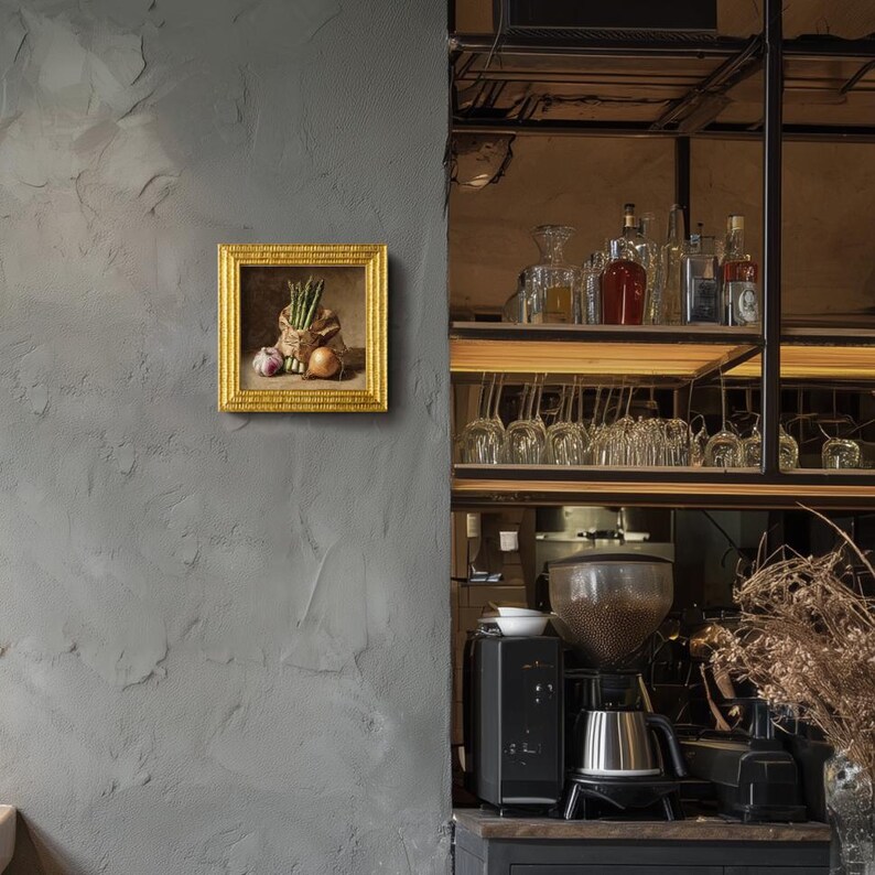 May include: A framed still life painting with a gold frame. The painting depicts a burlap sack filled with asparagus, an onion, and a garlic bulb. The painting is mounted on a textured gray wall, with a bar and coffee station in the background.