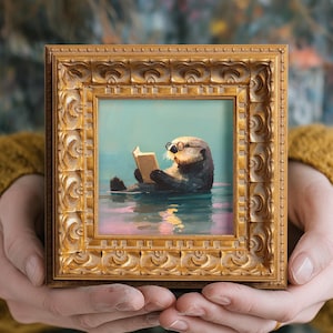 May include: A small square painting in an ornate gold frame depicting a sea otter wearing round glasses and reading a book while floating in calm water with soft pink and blue reflections on the surface of the water.