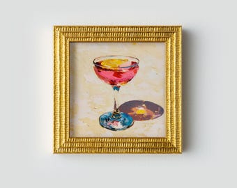 Peut inclure: Peinture encadrée d'un cocktail dans un verre coupe. La boisson est rose avec une garniture d'agrumes jaune. Le verre a une longue tige et une base bleue. Le cadre est doré.