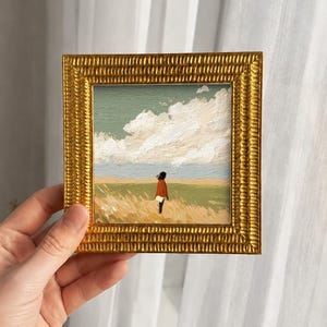 以下が含まれることがあります： 質感のあるゴールドのフレームに入った小さな正方形の油絵。くすんだ緑色の空の下、大きな白い雲が浮かぶ黄金色の野原に、赤いコートを着た人物が立っている様子が描かれており、目に見える筆致が特徴です。