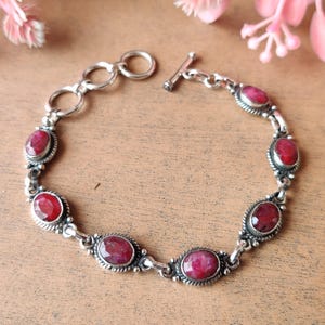 Puede incluir: Pulsera de plata con piedras preciosas ovaladas de color rojo intenso. La pulsera tiene un cierre de palanca y está compuesta por eslabones interconectados. Las piedras preciosas están engastadas en un marco de plata decorativo y con cuentas.