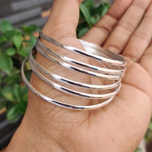 May include: A stack of six shiny silver bangles. The bracelets are smooth and round, reflecting light. The bangles are stacked on top of each other, creating a layered effect. The background is blurred, with green foliage visible.
