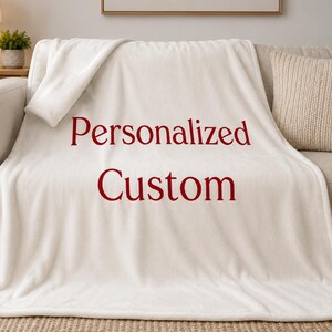 Könnte beinhalten: Eine weiche, cremeweiße Fleecedecke mit dem Text Personalized Custom, der in einer dunkelroten Serifenschrift in der Mitte aufgedruckt ist. Die Decke hat eine glatte, flauschige Textur und liegt über einem neutralfarbenen Sofa mit einer Ecke.