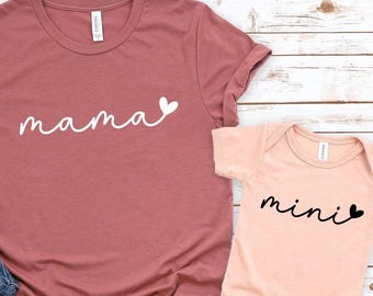 May include: A dusty rose t-shirt with the word "mama" in white script and a small white heart. Next to it is a peach-colored baby onesie with the word "mini" in black script and a black heart. Blue jeans and white sneakers are also in the image.