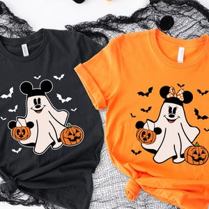 May include: A black t-shirt and an orange t-shirt, each featuring a graphic of a cartoon ghost wearing mouse ears and holding a jack-o'-lantern. The ghost on the black shirt has plain ears, while the ghost on the orange shirt wears a polka-dot bow.