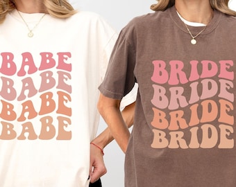May include: Two short-sleeve crewneck t-shirts. The ivory shirt features the word BABE repeated four times in a wavy, stacked pink and orange gradient font. The espresso shirt features the word BRIDE repeated four times in a similar wavy, stacked pink and orange gradient font.