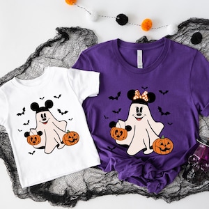 May include: A white t-shirt and a purple t-shirt, each featuring a graphic of a ghost with Mickey Mouse ears holding two jack-o'-lanterns, surrounded by black bats. The ghost on the purple shirt wears a polka-dot bow between the mouse ears.