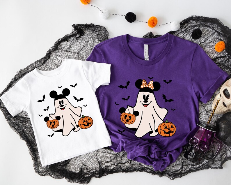 May include: A white t-shirt and a purple t-shirt, each featuring a graphic of a ghost with Mickey Mouse ears holding two jack-o'-lanterns, surrounded by black bats. The ghost on the purple shirt wears a polka-dot bow between the mouse ears.