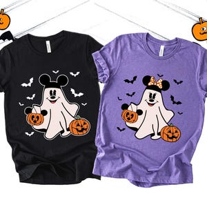 May include: A black t-shirt and a purple t-shirt, each featuring a white ghost graphic wearing mouse ears. The ghost on the black shirt wears plain black ears, while the ghost on the purple shirt wears polka-dot ears. Both hold orange jack-o'-lanterns.