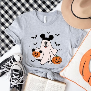 May include: A heather gray short-sleeve t-shirt featuring a graphic of a ghost wearing black mouse ears, holding a small orange pumpkin bucket, and standing next to a larger jack-o'-lantern, surrounded by several small black flying bats.