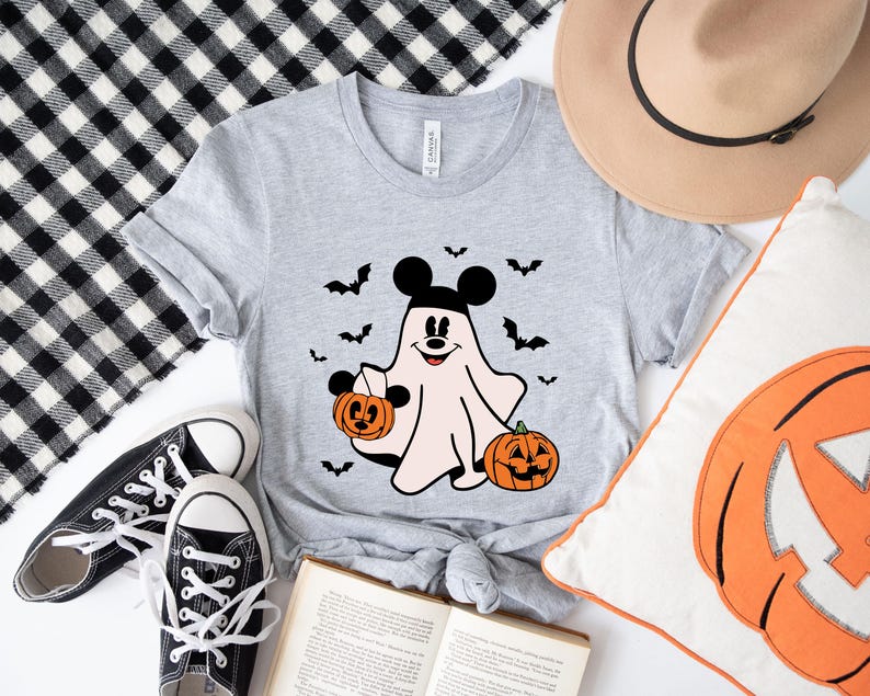 May include: A heather gray short-sleeve t-shirt featuring a graphic of a ghost wearing black mouse ears, holding a small orange pumpkin bucket, and standing next to a larger jack-o'-lantern, surrounded by several small black flying bats.
