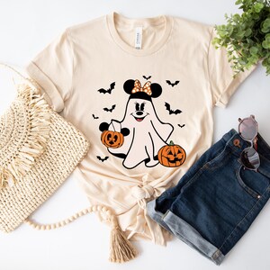 May include: A light beige short-sleeve t-shirt featuring a graphic of a ghost wearing mouse ears with a polka dot bow, holding a jack-o'-lantern bucket, accompanied by another jack-o'-lantern and several black bats printed on the front.