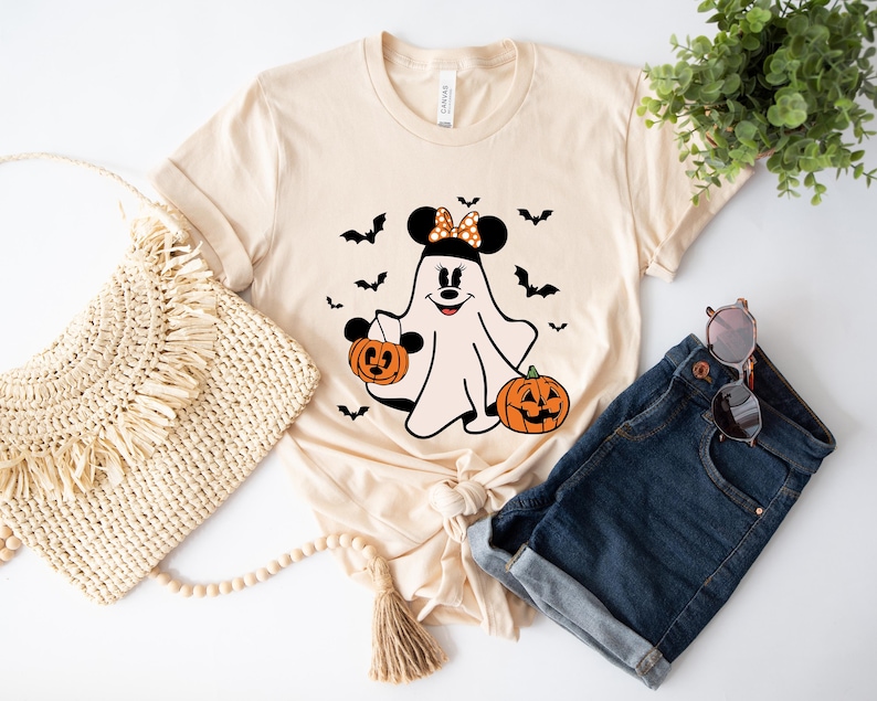 May include: A light beige short-sleeve t-shirt featuring a graphic of a ghost wearing mouse ears with a polka dot bow, holding a jack-o'-lantern bucket, accompanied by another jack-o'-lantern and several black bats printed on the front.
