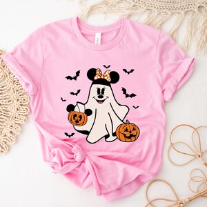 May include: A light pink short-sleeve t-shirt featuring a graphic of a white ghost character with black mouse ears, a polka-dot bow, and a smiling face. The ghost holds a small orange jack-o'-lantern, with another pumpkin and black bats nearby.