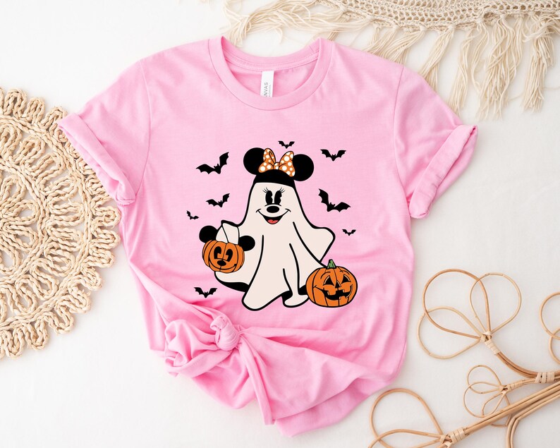 May include: A light pink short-sleeve t-shirt featuring a graphic of a white ghost character with black mouse ears, a polka-dot bow, and a smiling face. The ghost holds a small orange jack-o'-lantern, with another pumpkin and black bats nearby.