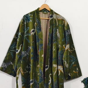 May include: A long, olive green velvet robe with a jungle-themed pattern of zebras, leopards, and tropical plants. The robe has a striped lining and a matching belt. It is hanging on a wooden hanger.