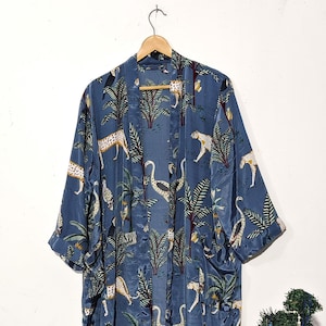May include: A long, flowing blue robe with a jungle-themed pattern. The robe features a repeating design of leopards, birds, and palm trees. The fabric has a silky sheen, and the robe hangs from a wooden hanger.
