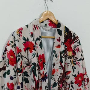 Puede incluir: Una bata de manga larga con base color crema y un estampado vibrante de flores y pájaros en tonos rojo, rosa, verde y azul oscuro. La prenda cuenta con un cuello ancho, un cinturón para anudar y un forro interior visible con rayas azules y blancas.